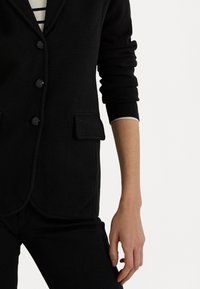 Černý blazer s propracovaným střihem, s klopovým límcem, pěti knoflíky a dvěma předními kapsami, vyrobený z texturované tkaniny.