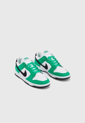 Paire de baskets basses Nike avec des panneaux verts, blancs et noirs et des lacets verts sur un fond clair uni.