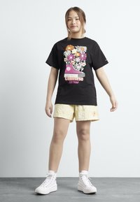 T-shirt noir avec graphique floral et design de baskets Converse roses. Shorts texturés jaunes. Baskets montantes blanches. Fond simple.