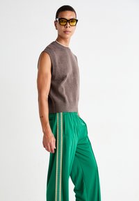 Top de punto marrón sin mangas combinado con pantalones verdes que tienen una franja lateral con un patrón de zigzag. La modelo lleva gafas con un tinte amarillo.