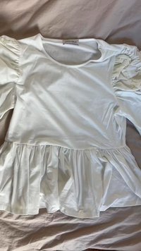 Bela bomba iz bombaža z okroglo ovratnikom, napihnjenimi rokavi in zategnjeno peplum obrobo. Nalepka nosi napis "ANNA FIELD." Gladka tekstura, sproščen dizajn.