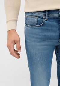 Jasno beżowy sweter o teksturowanej fakturze z prążkowanymi mankietami noszony na niebieskich, wyblakłych dżinsach z pięcioma kieszeniami i kontrastowym szwami.