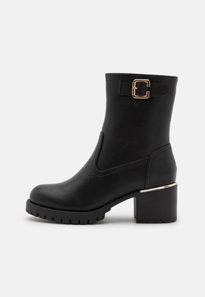 Botas de tobillo negras hechas de material sintético texturizado. Presentan un detalle de hebilla dorada, un tacón robusto y una suela de goma con tacos.