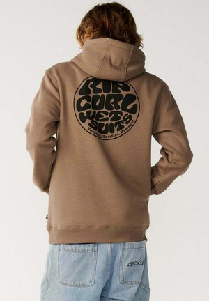 Persona que lleva una sudadera con capucha marrón de Rip Curl con un gran logo redondo en la espalda y vaqueros de mezclilla azul claro con un pequeño logo de Rip Curl en el bolsillo.
