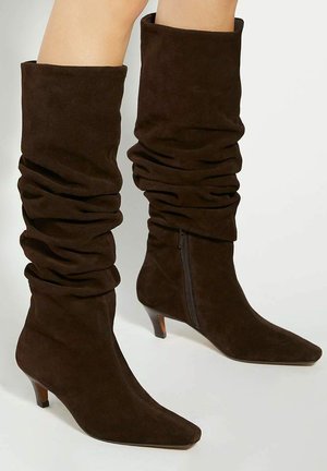 Brown suede knee-high boots with a slouchy design, pointed toe, and a small heel. Feature a zip closure on one side.