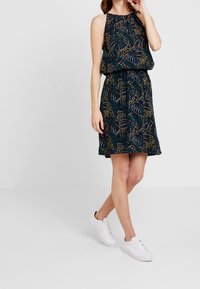 Robe à motif floral avec un col halter, une taille élastique et une jupe évasée. Présente un fond sombre avec des feuilles et des accents en or et vert.