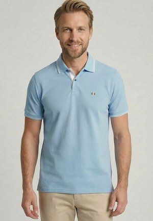 Lachende man met lichte baard, draagt een lichtblauw poloshirt en beige broek, staand tegen een eenvoudige grijze achtergrond.