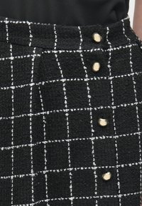 Fekete-fehér kockás szoknya texturált anyagból, négy arany gombbal az elején, fekete felsővel viselve.