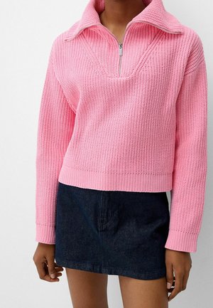 Pull en tricot rose avec un col zippé à quart, comportant un motif texturé. Porté par-dessus une jupe en denim foncé, mettant en avant des styles contrastés.