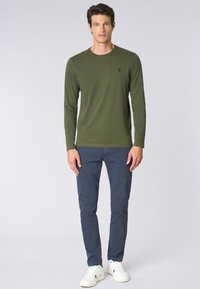 Camicia a maniche lunghe verde oliva con scollatura rotonda, abbinata a pantaloni slim-fit blu navy e sneakers bianche, con un logo discreto.