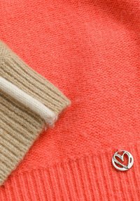 Close-up van koraalkleurige brei-stof met een beige en witte geribbelde manchet en een klein, zilverkleurig metalen hartvormig bedeltje ervoor genaaid.