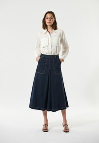Hvid skjorte med flæsekrave og brystlommer; mørkeblå denimnederdel med sidesnit og guldstikninger; sorte sandaler med stropper.