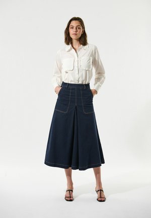 Chemise blanche à boutons avec un col froncé et des poches poitrine ; jupe en denim bleu foncé avec des fentes sur les côtés et des coutures dorées ; sandales noires à lanières.