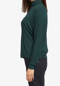 Maglione a maniche lunghe e collo alto di un verde scuro, realizzato in tessuto morbido e liscio. Presenta una vestibilità comoda e una texture a coste, abbinato a pantaloni neri.