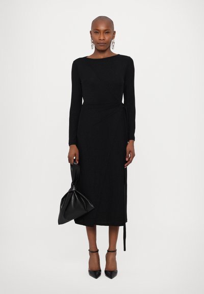 Vestido envolvente negro de manga larga en una tela suave, con escote barco y detalle de lazo en el costado. Combinado con unos tacones negros puntiagudos y un clutch.
