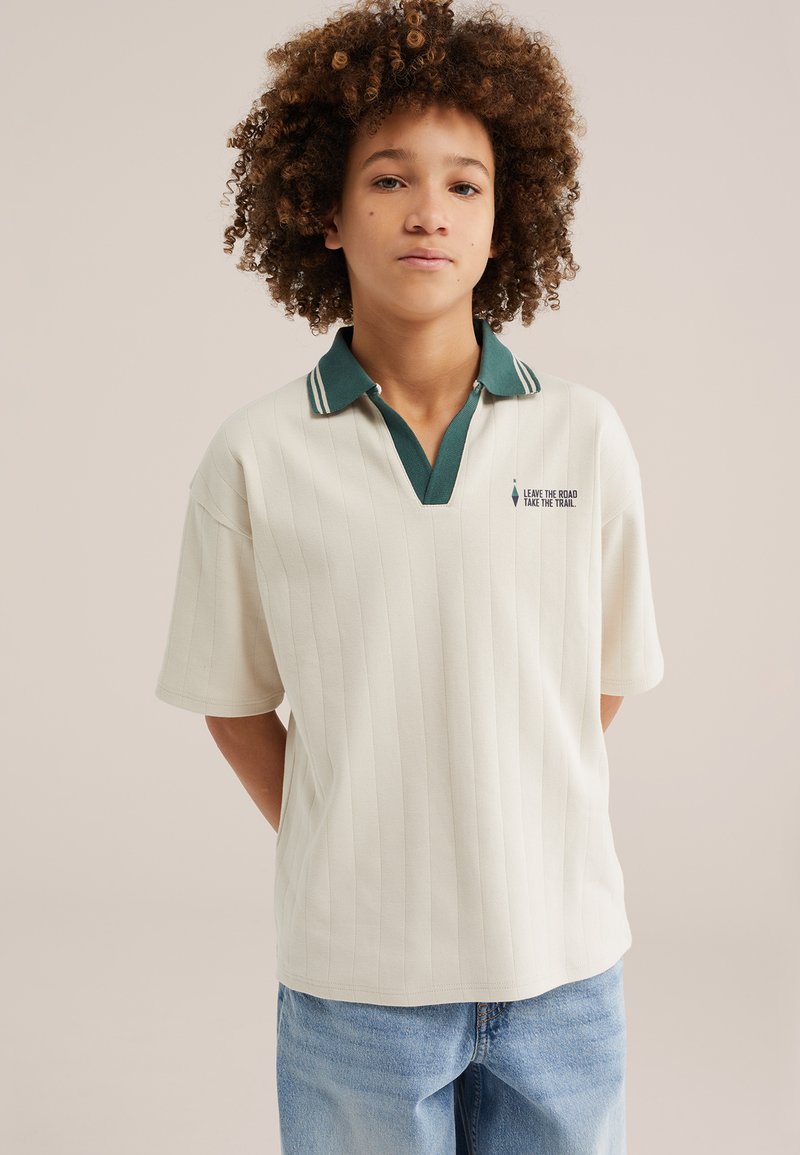 Adolescent aux cheveux bouclés portant un polo à manches courtes blanc avec un col vert foncé et un jean bleu, debout devant un fond uni.