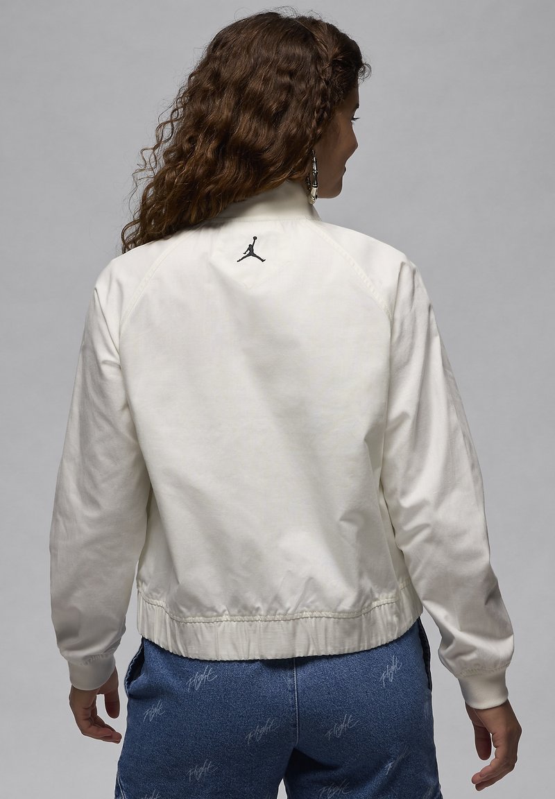 Veste bomber blanche avec un col haut, des poignets côtelés et un logo noir au dos. Tissu lisse avec une finition légèrement brillante.