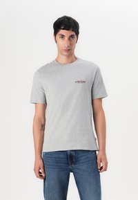 Tricou scurt gri din bumbac, cu guler rotund și un mic logo roșu "GUESS JEANS" pe față. Purtați cu blugi albaștri.