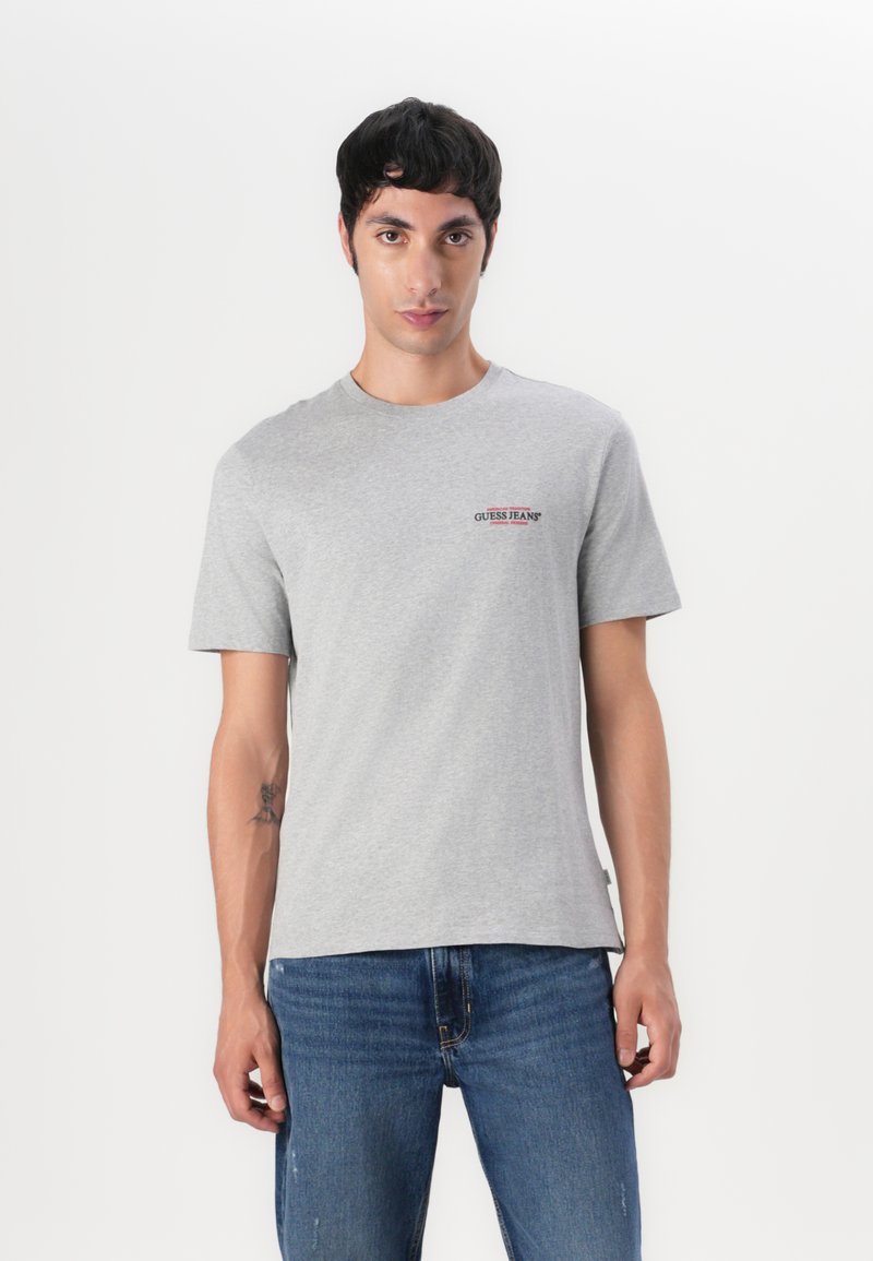 Tricou scurt gri din bumbac, cu guler rotund și un mic logo roșu "GUESS JEANS" pe față. Purtați cu blugi albaștri.