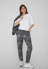 Graue Camouflage-Sweatpants mit einem Kordelzugbund, kombiniert mit einem weißen Kurzarm-T-Shirt und einer passenden Jacke, getragen mit weißen Sneakern.