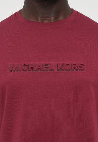 Bordeauxfarvet bomulds t-shirt med en forhøjet, tonet "MICHAEL KORS" logo på tværs af brystet. Korte ærmer og rund halsudskæring. Glat tekstur.