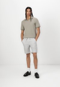 Kortermet, olivengrønn strikket polo skjorte med ribbestrikket krage, kombinert med lysegrå shorts med snor og svarte loafers. Hvite sokker er synlige.