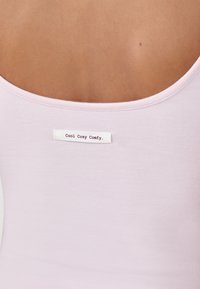 Jasnoróżowy top na ramiączkach z okrągłym dekoltem z tyłu. Zawiera białą metkę z napisem "Cool Cosy Comfy" wszytą z tyłu. Miękka, gładka faktura materiału.