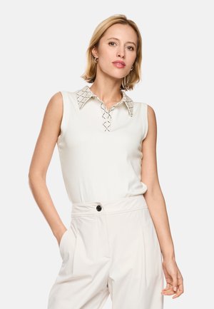 Woman with short blonde hair wears a sleeveless white blouse with a patterned collar and high-waisted white trousers, hand in pocket.