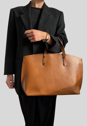 Borsa a tracolla in pelle marrone con due manici allungati, caratterizzata da una texture liscia e borchie circolari. Tenuta da una persona in un blazer nero.