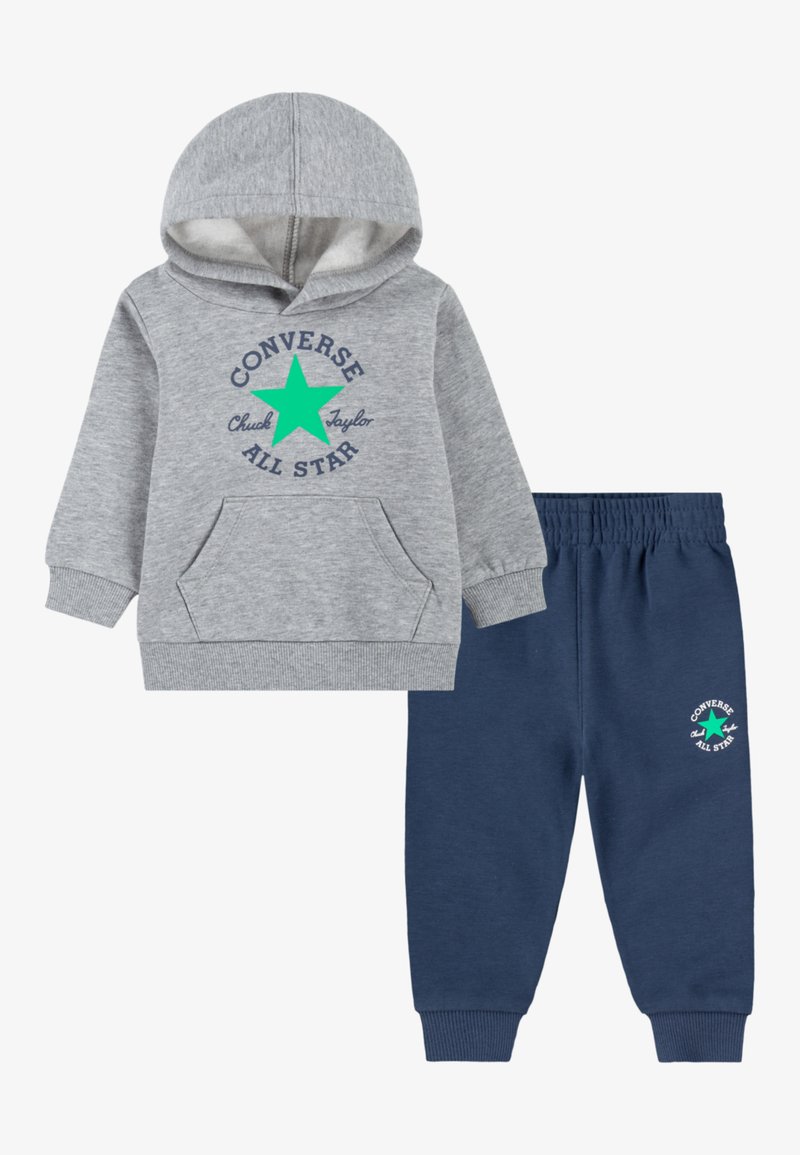 Siva kapuca z zadrgo z sprednjim žepom in zelenim logotipom zvezde, v kombinaciji z mornarsko modrimi trenirkami, ki imajo ustrezen logotip na levi nogi. Mehka tekstura tkanine.