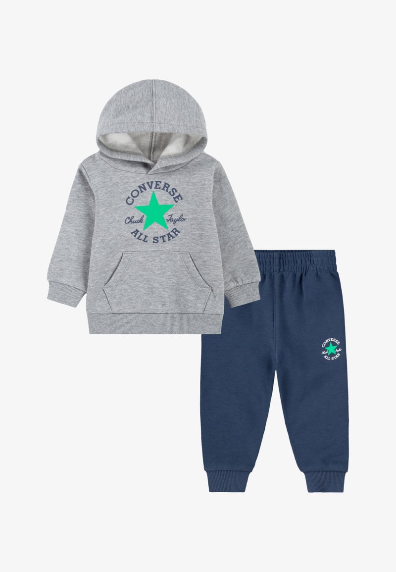 Siva kapuca z zadrgo z sprednjim žepom in zelenim logotipom zvezde, v kombinaciji z mornarsko modrimi trenirkami, ki imajo ustrezen logotip na levi nogi. Mehka tekstura tkanine.