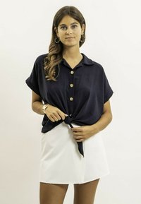 Chemise courte à manches courtes de couleur marine avec un col et un devant noué, ornée de boutons dorés. Assortie à une mini jupe blanche.