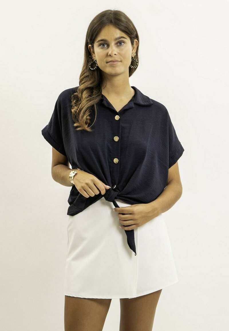 Chemise courte à manches courtes de couleur marine avec un col et un devant noué, ornée de boutons dorés. Assortie à une mini jupe blanche.
