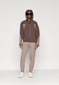 Καφέ μπουφάν με γκρι λεπτομέρειες, τσέπες με φερμουάρ και κουκούλα. Συνδυασμένο με μπεζ παντελόνι και ανοιχτόχρωμα αθλητικά παπούτσια. Μαύρα γυαλιά ηλίου.