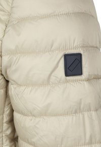 Beige gewatteerde jas met een gestikt patroon, voorzien van een stoffen patch met een blauw vierkant logo aan de linkerkant. Gladde textuur, lichtgewicht ontwerp.