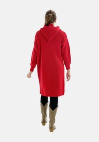 Vestido de sudadera de punto rojo con mangas largas, de largo hasta la rodilla, textura acanalada y cuello alto. Combinado con leggings negros y botas de vaquero beige.