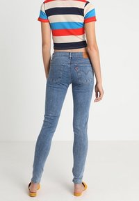Persona vestida con jeans ajustados azules y una camiseta de manga corta a rayas de colores, de pie con la espalda hacia el espectador sobre un fondo liso.
