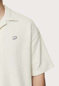 Camicia corta bianca sporca con una piccola logo ricamata "P" sul lato sinistro del petto, indossata da una persona su uno sfondo neutro.