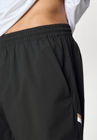 Črne športne kratke hlače z elastičnim pasom, z bočnimi žepi in kontrastnim vodoravnim trakom na spodnjem delu strani. Gladek tekstura.