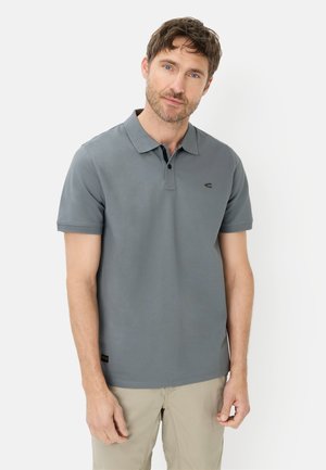 Man draagt een grijs poloshirt met korte mouwen en beige broek, staand tegen een effen witte achtergrond, naar voren gericht.