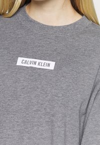 Sweat-shirt gris en matériau doux avec une étiquette rectangulaire blanche portant le texte "CALVIN KLEIN". Encolure arrondie et coupe décontractée.