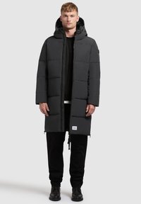 Padded czarny płaszcz z kapturem, z frontowym zamkiem błyskawicznym, kieszeniami po bokach i logo na dolnej krawędzi. Noszony na czarnej koszulce i spodniach.