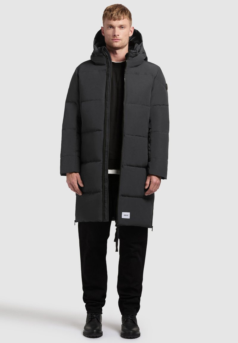 Padded čierny kabát s kapucňou, ktorý má predný zips, bočné vrecká a logo na leme. Nosený cez čiernu košeľu a nohavice.