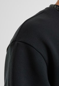 Schwarzer Sweatshirt mit weicher Textur, abgerundeten Schultern und sichtbaren Nähdetails; besitzt einen Kette-Akzent am Ausschnitt.