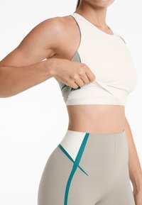 Lättvikts, figurnära sportbh i beige med hög hals och teal-detaljer, kombinerad med högmidjade grå tights med liknande detaljer.