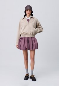 Giacca beige con zip e orlo elastico, abbinata a una gonna plissettata a quadri rossi e bianchi. Calze grigie e scarpe marroni con dettagli dorati.
