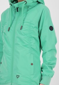 Femme portant une veste à capuche vert menthe avec cordons de serrage, fermeture éclair, poches latérales, poignets côtelés, et un petit emblème en forme de cœur près de l'ourlet.