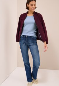 Femme portant une chemise bleu clair, une veste bomber bordeaux, un jean bleu et des baskets beiges, se tenant dans un coin aux tons neutres.