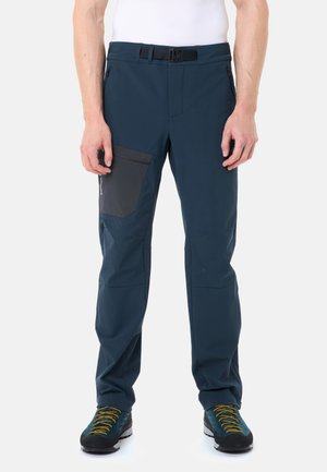 Uomo che indossa pantaloni outdoor blu scuro con tasca sulla coscia con zip nera e scarpe da trekking blu con lacci gialli, in piedi su sfondo bianco.