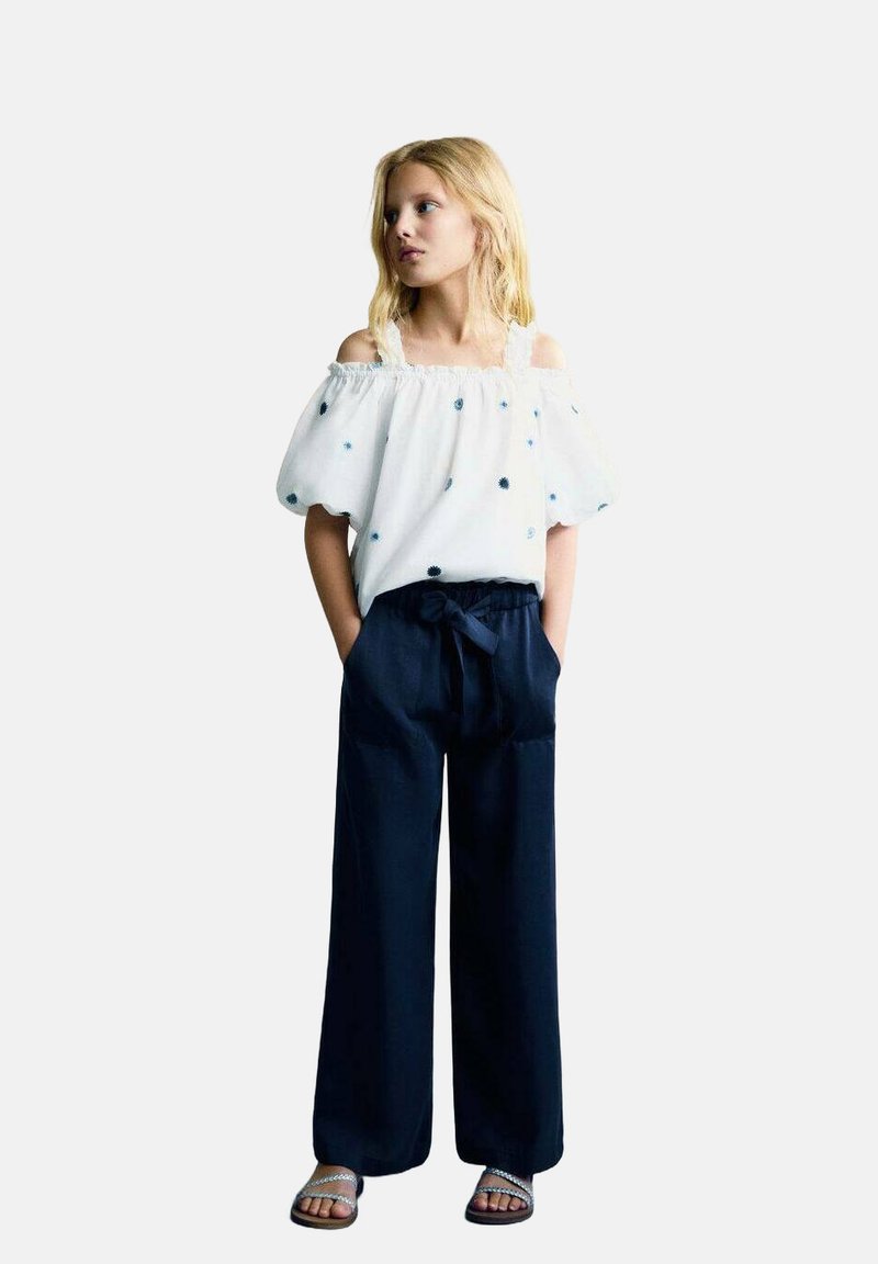 Blusa bianca a spalle scoperte con maniche a sbuffo e pois blu ricamati. Pantaloni navy a gamba larga con vita annodata, indossati con sandali bassi.